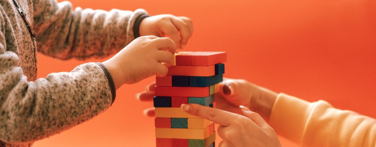 A child and adult play with colorful wooden blocks against an orange backdrop, emphasizing learning and creativity.